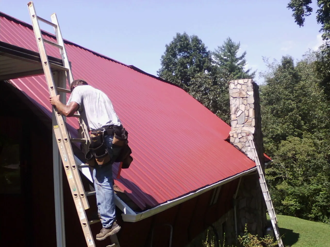 Roofer on a ladder beside a red metal roof and stone chimney — Soffit & Fascia Repair by Roofing Brothers in Round Rock